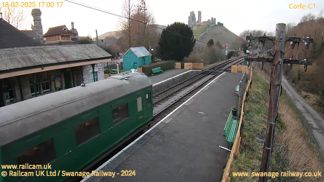 A green train car is parked at a railway station platform. The platform has several green benches and some trees in the background. In the distance, a castle is visible on a hilltop. The sky has a light hue, indicating it may be early evening. A wooden fence borders the platform, and there are power lines along the edge of the platform.