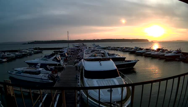 A marina at sunset, featuring numerous boats docked in calm water. The sky is overcast with hints of orange and yellow from the setting sun on the horizon. In the foreground, a large white boat is visible, and there are several smaller boats lined up along the dock. The scene conveys a tranquil evening atmosphere by the water.