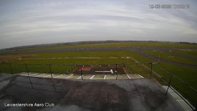 A view from a webcam at Leicestershire Aero Club shows a grassy airfield with a paved runway in the foreground. A safety railing is visible at the bottom of the image. There is a light cloud cover in the sky, and the scene appears to be overcast. The airfield features a few scattered markings on the runway, and a windsock on a pole indicates wind direction. The date and time stamp in the corner reads 18-02-2025 at 17:00:42.