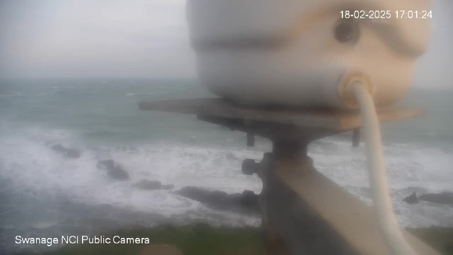 A blurred image of a coastal scene taken from a webcam. In the foreground, there is a white cylindrical object mounted on a post, connected by a cable. The background features a turbulent sea, with waves crashing against rocky formations. The overall image appears hazy, obscuring details of the landscape. The timestamp indicates the image was taken on February 18, 2025, at 17:01:24.