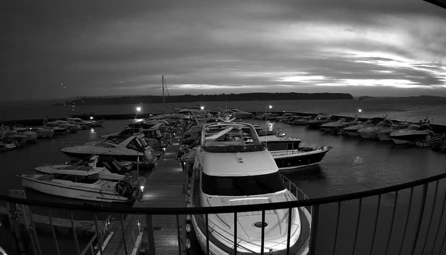 A black and white image shows a marina filled with various boats docked at a pier. The scene is illuminated by lights from some of the boats, reflecting softly on the water. The sky is overcast, with dark clouds suggesting an impending storm, and the silhouette of land can be seen in the background.