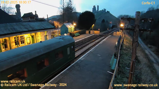 A dimly lit train station with a green railway carriage departing to the left. The platform features a wooden fence, benches, and a station building with large windows illuminated from within. In the background, a hill is visible with ruins on top, and trees are scattered around. The sky is transitioning to twilight, with soft blue and orange hues, and electrical poles are seen on the right side of the image.