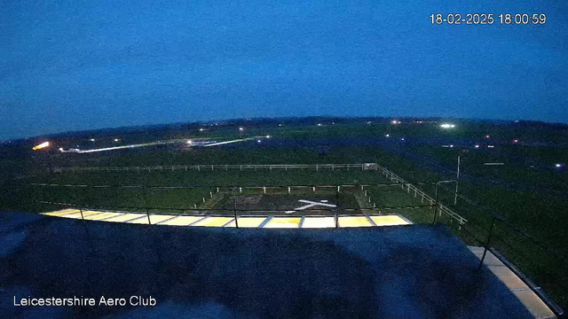 A dimly lit scene of an aerodrome at twilight, featuring a grassy runway and a nearby helipad marked with a white cross. The sky is blue-grey with fading light, and various lights outline the runway. Fences are visible in the background, with a few distant lights suggesting vehicles on or near the tarmac. The foreground includes a partial structure with illuminated panels.