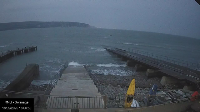 A cloudy and gray scene at the coast, showing choppy waves hitting a concrete pier. To the left, a set of stairs leads down toward the water. In the foreground, a yellow kayak and a blue kayak are visible near a railing. The distant shoreline features cliffs, partially obscured by fog or haze. The date and time are displayed in the corner.