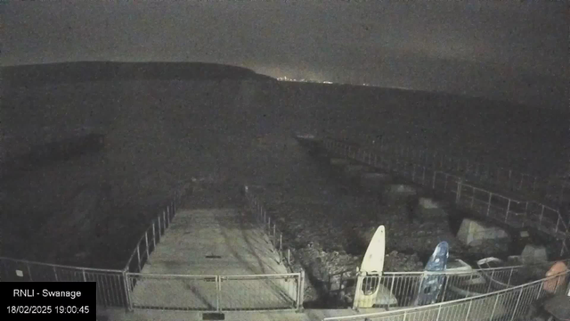 A dimly lit scene at a coastal area, featuring a stone pier with a metal railing. On the pier, there are two surfboards, one pale yellow and one blue, propped against the railing. The water is visible but dark, extending to the horizon where faint distant lights can be seen. The landscape has a hilly outline in the background, suggesting a rugged coastline. The overall atmosphere is quiet and somewhat serene, with a nighttime setting.