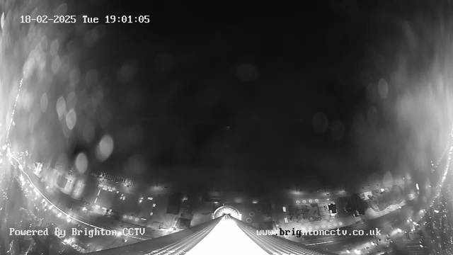 A black and white webcam image featuring a view from a high vantage point. The scene is mostly dark with scattered light reflections, possibly from rain on the lens. Below, a brightly lit street is visible, lined with vehicles and light sources. The date and time are displayed in the top left corner, indicating it is February 18, 2025, at 19:01:05. The bottom left corner contains text reading "Powered By Brighton CCTV."