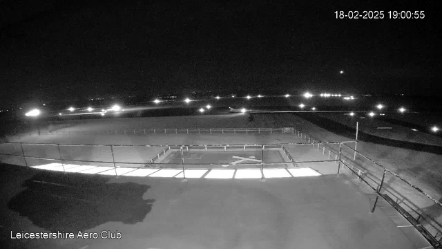 A nighttime view of an airfield with low visibility. The scene is dark with scattered lights illuminating parts of the area. A rectangular runway is visible in the center, marked with a white cross. There are fences along the perimeter, and additional lights in the background suggest other structures or activities nearby. The sky is mostly dark, with a faint light in the distance.