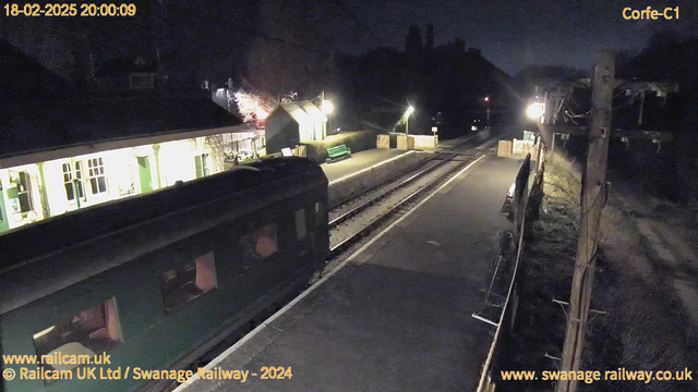 A dimly lit railway station at night, with a green train car in the foreground. The platform has a few benches, one of them green, and there are wooden fencing panels surrounding the area. In the background, a small building with bright lights and windows is visible, along with some trees. The railway tracks run through the scene, illuminated by a distant light source.