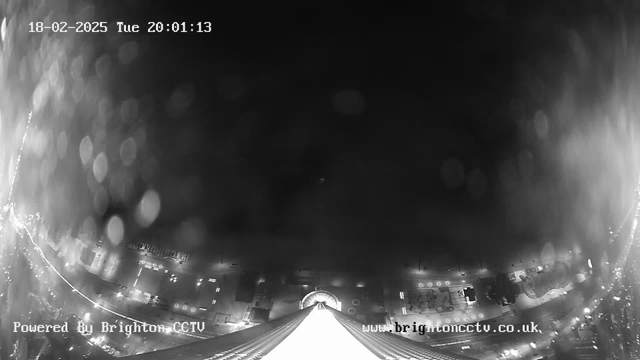 A black and white image from a webcam shows a view of a city at night, partially obscured by raindrops on the lens. The foreground features a wide white structure leading into the distance, gradually tapering off. Below, there are faint outlines of streetlights and vehicles, which create a sense of urban life. The top of the image displays the date and time in white text, indicating February 18, 2025, at 20:01:13. The corners have text indicating the image is powered by Brighton CCTV and providing a website link.