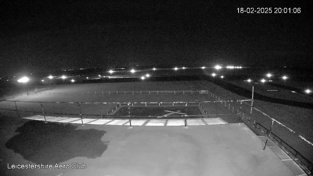 A nighttime view from a webcam showing an open area. The foreground contains a flat surface with a large rectangular section, partially illuminated. There are several lights in the distance creating a faint glow, and a fence runs along the edge of the area. The overall scene is dark, with the faint outlines of a field in the background and a few distant lights scattered across the horizon.