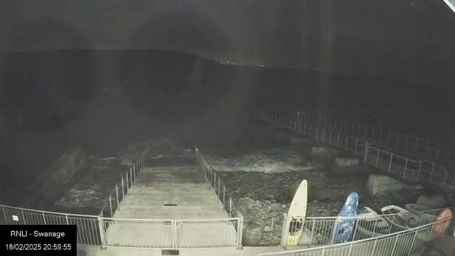 A dimly lit scene showing a rocky shoreline at night. In the foreground, there is a set of concrete steps leading down to the water, enclosed by a metal railing. To the right, several boats and kayaks with bright colors, including yellow and blue, are resting on the ground. The ocean appears dark and choppy, with visible waves, and a distant shoreline can be seen in the background, slightly illuminated. The time and date are displayed in the bottom left corner of the image.