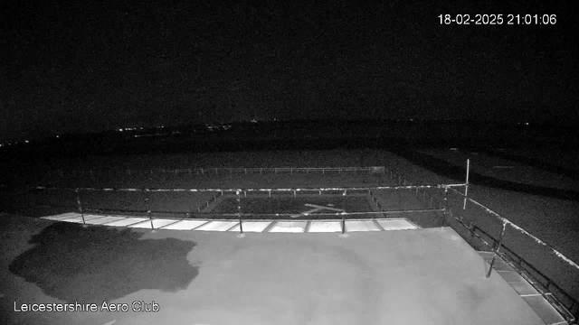 A dark, nighttime scene showing a flat surface with a light patch on the left side, likely reflecting nearby lighting. In the background, faint lights are visible in the distance, suggesting a landscape with low visibility. There is a metal railing in the foreground, and an open area that appears to be part of an airstrip or runway. Text in the bottom left corner indicates "Leicestershire Aero Club" and there is a timestamp in the top right corner.