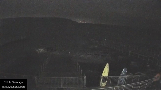 A dark scene captured from a webcam showing a shoreline with a faint outline of hills in the background. There are two kayaks, one yellow and one blue, positioned by a fenced area. The water appears calm, and the visibility is low, suggesting nighttime conditions.