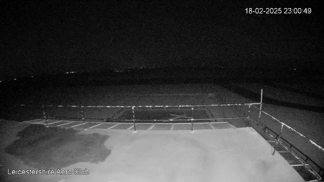 A dark nighttime scene captured from a webcam at the Leicestershire Aero Club. The image shows a flat surface in the foreground, possibly a landing strip or tarmac, with a railing along the edge. In the background, faint lights can be seen at a distance, suggesting the presence of a horizon or distant structures. The overall atmosphere is quiet and empty, typical of nighttime with minimal visibility. The timestamp indicates the image was taken on February 18, 2025, at 23:00:49.