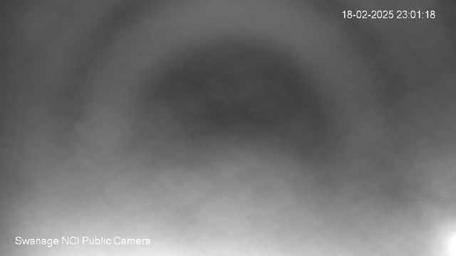 A grayscale image of an unclear, hazy scene, with a gradient of darker shades at the top transitioning to lighter shades at the bottom. The bottom left corner includes the text "Swanage NCI Public Camera," while the top right corner displays a timestamp. The overall visibility is low, with no distinct shapes or objects present.