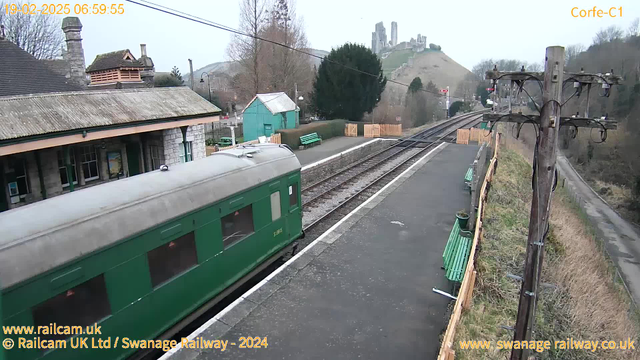 The image shows a scenic railway station platform with a green train car gradually moving away from the camera. In the background, there are several empty benches and a wooden fence on the left side of the platform. Beyond the platforms, a railway track extends into the distance. On the right, a wooden utility pole displays wires and attachments. In the background, a hill rises with ruins that appear to be a castle or fortification on its summit. The sky is overcast, creating a muted atmosphere.