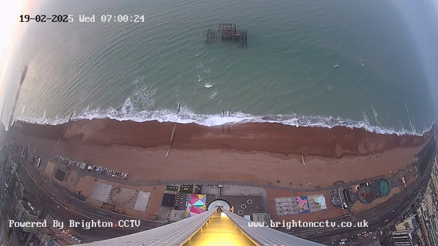 Aerial view of a beach at dawn, featuring a sandy shoreline meeting the waves of the sea. Off to the left, a pier extends over the water, while in the center, remnants of a wooden structure are visible just above the waterline. The beach is lined with colorful structures and a circular layout of pathways. The scene is illuminated by the soft light of the early morning. The date and time are displayed at the top of the image.