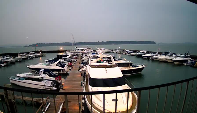 A view of a marina filled with various boats docked in a calm body of water. The scene is overcast, with a gray sky and a hint of fog. Several boats, mostly white and blue, are aligned along the wooden dock. In the distance, there is a hazy shoreline with trees. Some boats are equipped with visible covers and equipment on their decks. A few lights hint at activity on the dock, but overall, the atmosphere is quiet and serene.