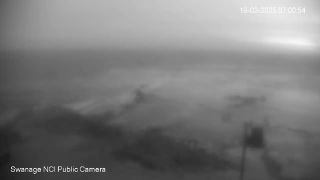 A black and white image of a coastal scene viewed from a webcam. The horizon is faintly visible, blending into a cloudy sky. The water appears calm with soft ripples, and the foreground shows indistinct shapes, possibly debris or rocks. A pole or post with a flag is slightly visible on the left side of the image. The overall atmosphere is foggy and muted, suggesting low visibility conditions. The time stamp indicates it was taken early in the morning.