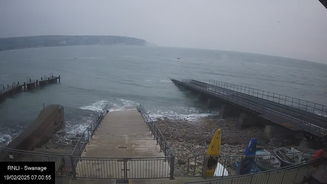 A view of a rocky shoreline and the ocean under an overcast sky. In the foreground, a set of concrete steps leads down to the water, bordered by railings. To the right, there are several colorful kayaks stacked against a railing. A wooden pier extends into the water, visible on the left side of the image. The water is choppy with small waves, and the distant coastline is partially obscured by fog.