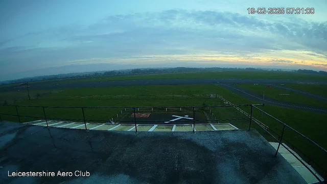 A view from a rooftop at the Leicestershire Aero Club. The scene shows a grassy airfield with a gray runway extending into the distance. A large cross shape, likely a landing marker, is visible in the foreground. The sky is mostly cloudy, with soft blue and orange hues indicating early morning light. Fencing borders the airfield and a few structures are discernible along the horizon.