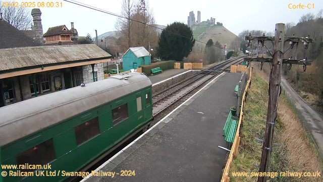 A green train carriage is parked at a railway station. The station features a stone building with a peaked roof and wooden accents. There are green benches along the platform and a wooden fence in the background. In the distance, a hill rises with ruins of a castle at the top. The sky is overcast, and the scene is predominantly gray and green. A utility pole with wires is situated on the right side of the image.