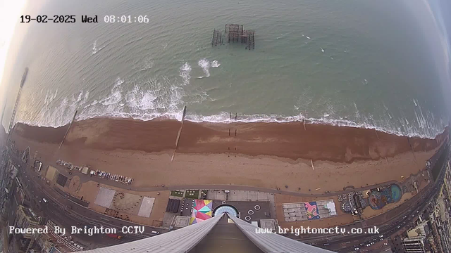 A high-angle view of a beach scene at dawn. The sandy beach is visible along the bottom of the image, with gentle waves lapping at the shore. A pier extends out into the water, with some structures visible above the surface. To the right, there are amusement rides and buildings, indicating a lively seaside area. The sky is lightening with the early morning light, and the date and time are displayed in the top left corner.