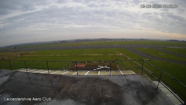 A view of an open airfield taken from a rooftop. The ground is mostly covered in green grass, with some patches of brown. There are several white markings on the pavement denoting a runway area. A fence surrounds a section of the grass, and in the distance, there are faint outlines of trees and hills under a cloudy sky. The scene is bright but overcast, indicating a dull day. The time is recorded as 08:00:34 on February 19, 2025.