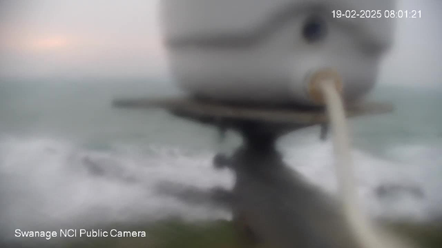 A blurred image depicting a coastal scene where the ocean meets a grey sky. In the foreground, there is an indistinct structure resembling a round container with a hose attached. Waves are visible in the background, suggesting a windy or stormy atmosphere. The overall image is hazy, contributing to a sense of overcast conditions.