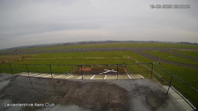 A view from a webcam situated at the Leicestershire Aero Club, looking over a grassy airfield. In the foreground, there is a railing and some white markings on the ground. The airfield is mostly clear, with a small section of paved runway visible, and green grass stretching out in the distance. The sky is overcast with gray clouds, and there are hills faintly visible in the background.