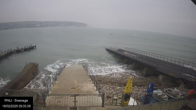 A view of a rocky shoreline and gentle waves lapping against the edge of a concrete ramp leading down to the water. To the left, a small stone structure juts into the sea, while to the right, a wooden pier extends into the water. Several colorful kayaks, including a yellow one, are positioned on the ground near the ramp. The sky is overcast, blending with the muted colors of the water and surrounding landscape.