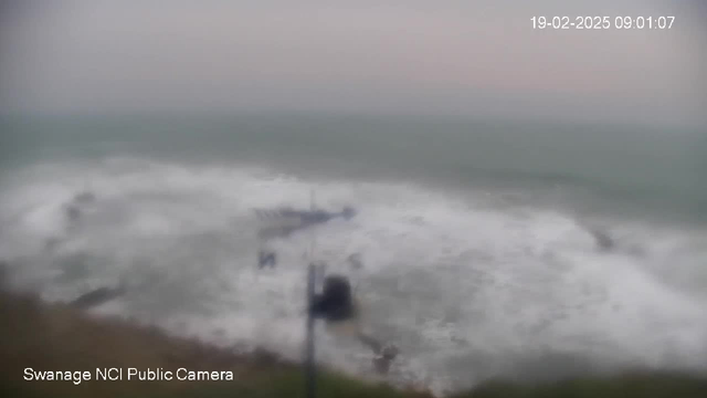 A blurry image of rough ocean waves crashing onto a rocky shore, under a grayish sky. The scene depicts a coastal area, with indistinct objects partially visible, possibly indicating structures near the water. The time is noted as 09:01:07 on February 19, 2025.