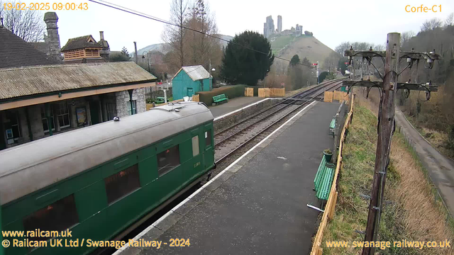 A green railway carriage is visible on the left side of the image, with its side windows reflecting surroundings. The platform is made of grey stone and has several wooden benches painted green. In the background, there is a small blue building and lush trees. A hill rises in the distance, topped with the ruins of a castle. The sky is overcast, indicating a cloudy day. There are railroad tracks running parallel to the platform, leading into the distance.