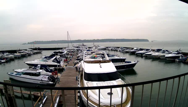 A marina filled with various boats docked in the water. There are several white and blue yachts visible, some with colorful buoys on their decks. The sky is overcast with a gray hue. In the distance, a shoreline can be seen with trees lining the edge, and gentle waves are present on the water's surface. A wooden dock is visible in the foreground, providing access to the boats.