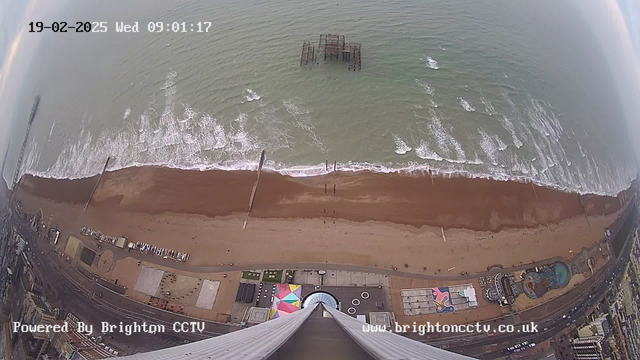 Aerial view of a beach with brown sand and gentle waves lapping at the shore. In the distance, a pier extends over the water. There are scattered objects on the beach and a colorful patch of land, possibly a fairground or amusement area. A portion of the shoreline appears developed, featuring buildings and structures along the coast. Above, the sky is mostly clear, with hints of clouds. The image is timestamped 19-02-2025, 09:01:17.