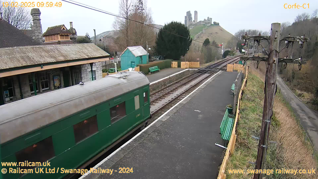 A green train carriage is partially visible on the left side of the image, with its door facing the platform. The platform is made of gray paving stones and includes several green benches along the edge. In the background, there is a small building with a peaked roof, and behind it, a green shed. A wooden fence runs along the right side of the platform, and a power pole with wires is positioned at the edge. In the distance, a hill features the ruins of a castle. The sky is overcast, and the scene gives a quiet, rural atmosphere.