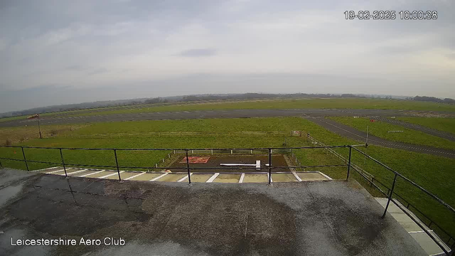 A view from a webcam positioned on a building, overlooking a grassy airfield. In the foreground, there is a railing and a section of the building's roof. The airfield is mostly empty, with several runways marked by lines and a few scattered objects. There is a windsock indicating the wind direction near the edge of the airfield. The sky is overcast, with a grayish tint. The date and time displayed in the corner indicate it is February 19, 2025, at 10:00 AM.