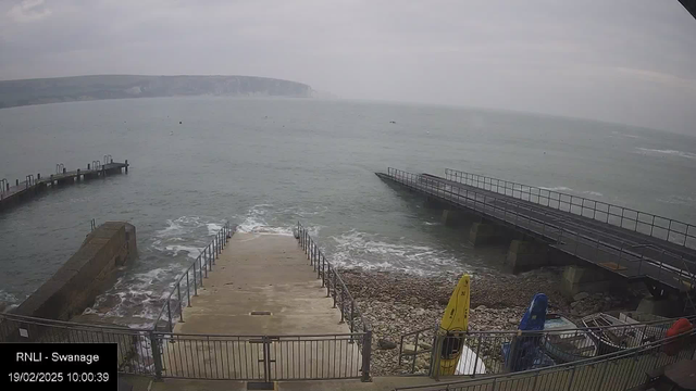 A view of a seaside location featuring a concrete ramp leading down to the water, with metal railings on either side. The ramp is partially submerged in shallow waves. In the background, there are two piers extending into the sea. The sky is overcast, and along the water's edge, there are smooth stones. Various colored kayaks, including yellow and blue, are parked on the beach to the right. The distant shoreline features a steep, rocky cliff.