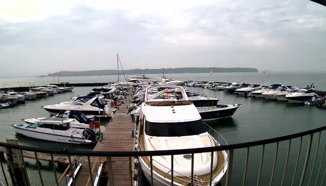 A view of a marina filled with various yachts and boats docked along a wooden pier. The scene is set against a cloudy sky, with water reflecting the boats and the shoreline in the background. There are multiple boats of different sizes and colors, some with covers, moored in neat rows. The atmosphere appears calm and serene.