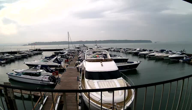 A view of a marina on a cloudy day, with several boats docked in calm waters. The scene includes a variety of boats, most of which are white and blue, and a wooden pier extending into the water. The background shows a distant shoreline and hills. The atmosphere appears gray and overcast.