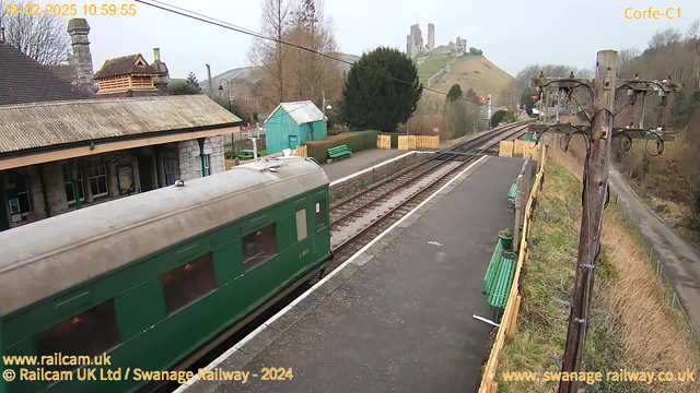 An outdoor scene at a railway station with a green vintage train on the left side of the image. The train is parked on the tracks, which run parallel to the platform. The platform features several green benches and a stone station building with a thatched roof, situated in the background. In the further distance, a hillside is visible with ruins of a castle and trees scattered around. Power lines run along the edge of the platform. The overcast sky gives a muted appearance to the overall scene.