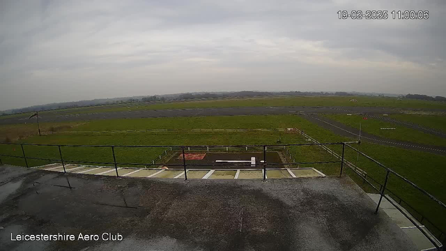 A wide view of an open grassy area under a cloudy sky, taken from a balcony or elevated platform. In the foreground, a railing outlines the edge of the platform. Below, a flat area with a white marking and a small red patch is visible. The background features a runway, with low rolling hills in the distance. The image is timestamped with the date and time.