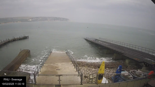 A cloudy sky looms over a calm sea with gentle waves. In the foreground, a set of concrete steps leads down to the water, flanked by a metal railing. To the right, a wooden pier extends into the sea, while another smaller structure juts out on the left side, with several brightly colored kayaks—yellow and blue—stored along the edge. The shoreline features pebbles and rocky areas, and some distant cliffs can be seen in the background across the water. The time stamp indicates the image was captured at 11:00 on February 19, 2025.