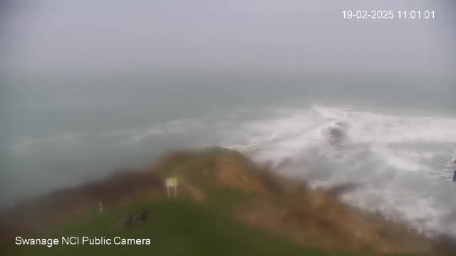 A foggy coastal scene is visible, with waves crashing against rocky shoreline. In the foreground, there is a patch of grass and a signpost, while the ocean appears choppy with white frothy waves. The atmosphere is overcast and the visibility is low. The date and time are displayed in the top right corner.