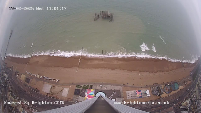 Aerial view of a sandy beach meeting the ocean. The coastline features waves crashing onto the shore, with a wooden pier extending into the water. In the foreground, various structures and amusement park setups are visible, along with a colorful pattern on the ground. The sky is overcast, and the image shows the time and date in the top left corner.