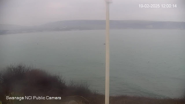 The image shows a coastal scene captured from a webcam at Swanage. The view features calm, gray water with a few small boats visible on the surface. The shoreline is slightly obscured by fog or overcast conditions, and there are low hills or cliffs in the background. In the foreground, there is some vegetation and a pole, possibly part of the webcam setup. The overall atmosphere is muted and tranquil.