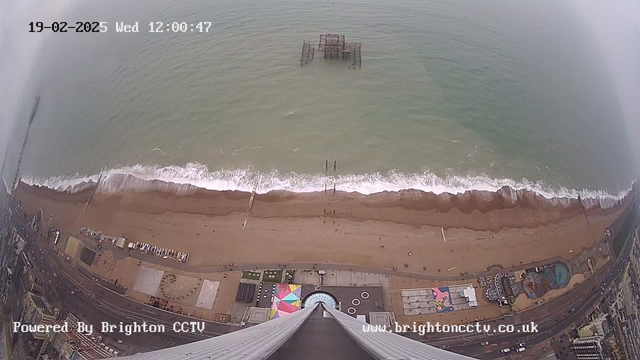 A high-angle view of a sandy beach with waves crashing onto the shore. The image shows a partially visible amusement pier extending into the sea with several wooden posts. Along the beach, there are colorful structures and a series of attractions. The sea appears calm with a slight mist or fog in the background. The date and time are displayed in the top left corner.