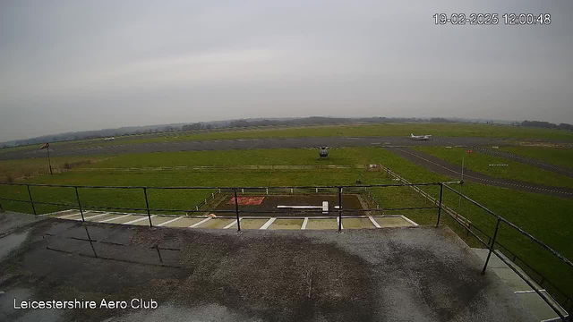 A view from a webcam at an aerodrome. The foreground shows a flat rooftop with a railing and some water on the surface. In the background, there is a grassy area with a paved runway and another small aircraft moving to the right. The sky is gray and overcast, indicating possibly cloudy weather.