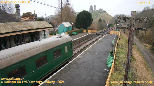 A green train car is positioned on a railway track at a station. The station features a stone building with a sloped roof, and behind it is a wooden shed painted light blue. Several green benches are along the platform. In the background, a hill rises steeply, topped with the ruins of an old castle. The sky is overcast, giving the scene a muted light. Electrical poles with wires are on the right side of the image.