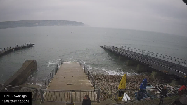 A cloudy day at the coast with a view of the sea. In the foreground, a set of wide, concrete steps leads down to the water, surrounded by a metal railing. To the left, there is a rocky area by the water's edge. On the right, two small boats are visible, one yellow and one blue, alongside a few other boats. In the background, a wooden pier extends into the sea, and a steep, grassy cliff rises in the distance. The atmosphere appears calm and quiet.